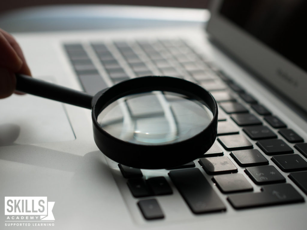 Person using a magnifying glass to look closely at a laptop. Start a successful career with our Forensic Investigations Courses.