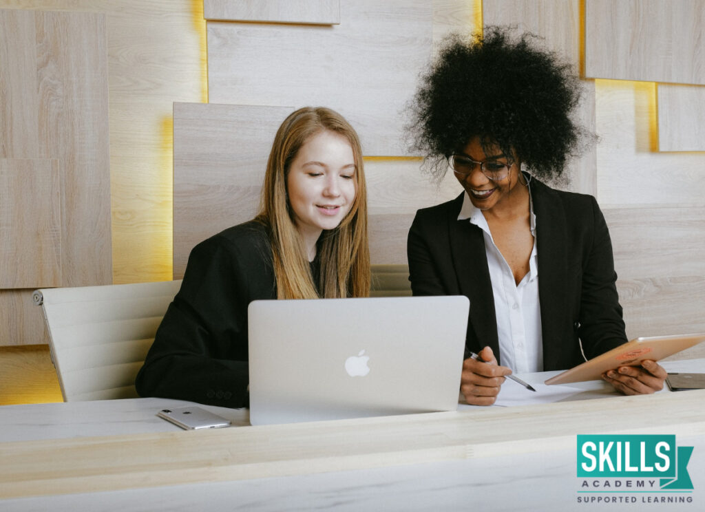 Secretary helping her colleague complete admin duties on a laptop. Improve your skills with our Secretarial Courses