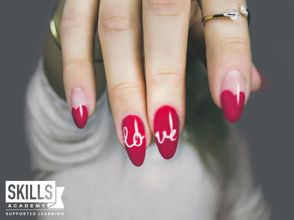 Woman's hand with red nail polish on and the word love written on her index and middle fingers. Lean naill application skills with us.