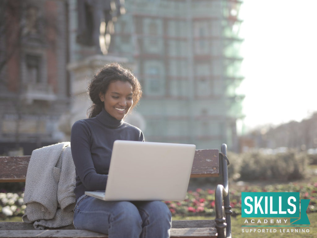 A young student sitting on a park bench studying on her Distance Learning course on her laptop.