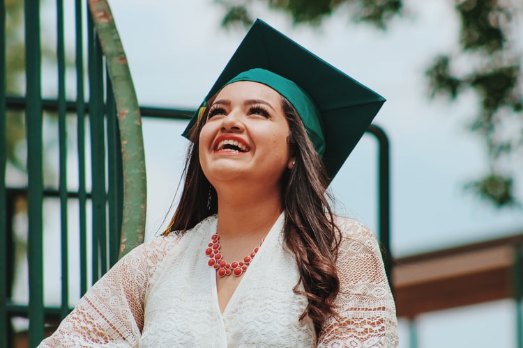 A young girl with a while dress, orange necklace and her green graduation cap on is looking up at the sky smiling happily because she took our advice for Job Seekers