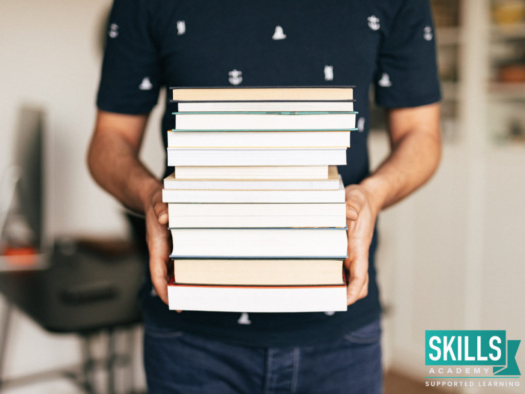 Student holding a stack of textbooks that contains Subjects Required for Business Management. Learn what they are by signing up for our courses.
