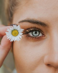A woman's side face adorned with beauty products. She has grey blue eyes and is carrying a flower next to her eye