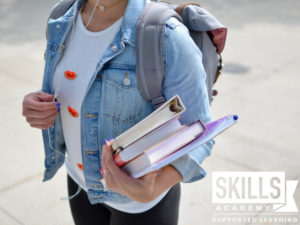 A college student in a denim jacket and white tshirt carrying books on her left arm and a carrying a backpack on her bak