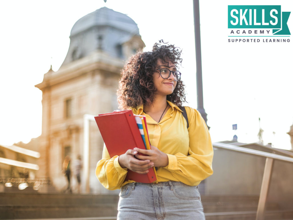 A young woman with a stack of books in her hands standing in front of a college. Make the right choice when choosing to study. Here are Things to Consider When Choosing a College.
