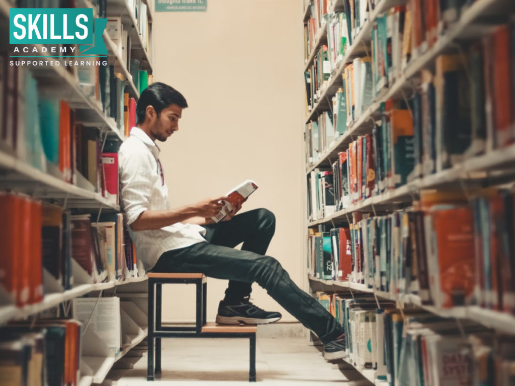 A young man who found out what subjects are needed to study financial management -reading a book in the library wearing a blue jeans and white sheet.