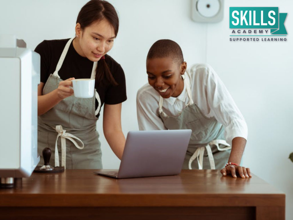 Two young women making use of their Basic Computer Skills to manage their coffee shop. Find out what Basic Computer Skills for a Successful Career are needed right here!