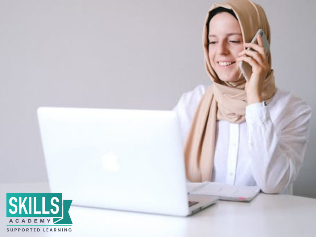 A young girl wearing a white shirt and brown hijab, sitting in front of her laptop, on the phone with a course expert speaking about Tips on Getting Into the Beauty Industry.