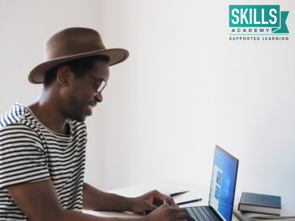A student with a black and white striped shirt on and a brown hat, sitting at his desk finding out how to get a bursary on his laptop.