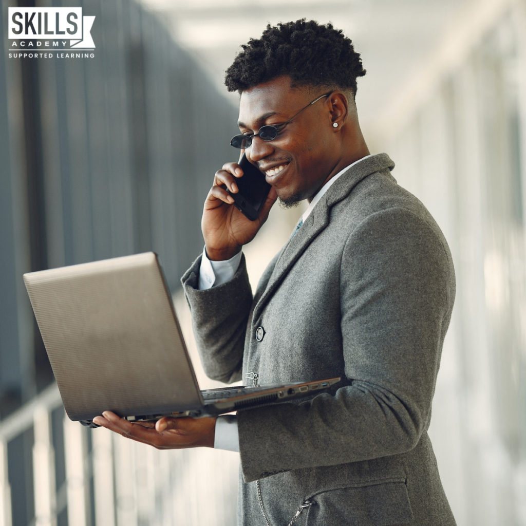 Man wearing a suit and sunglasses smiles while looking at a laptop and talking on a cellphone. Learn the skills you need with our Courses That Will Improve Your Leadership Skills.