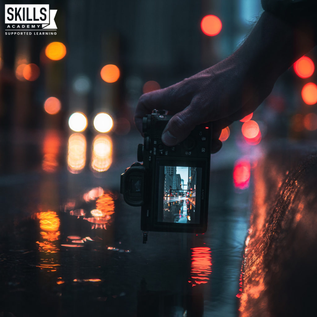 Man holding a camera with lights shining on a busy street. Learn how to take images like these with our Career Tips for Photography Enthusiasts.