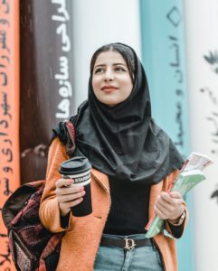 A student wearing a hijab standing with her books in her hand and a coffee cup in the other. Managing Student Loans and enjoying studying.