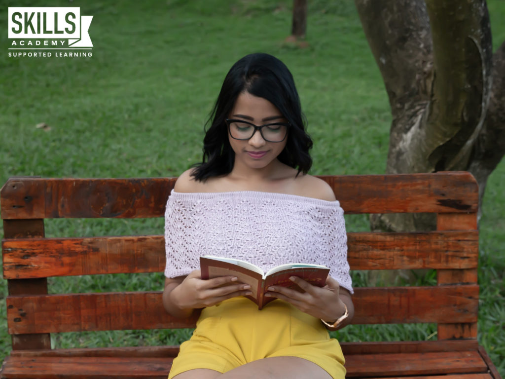 Girl wearing a white shirt and yellow shorts is sitting on a bench reading in a park. You can also studying from anywhere when you register at our distance learning college.