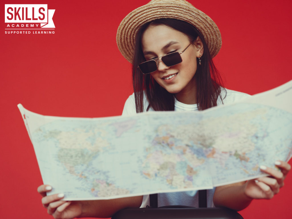 Woman looking at a map standing in front of a red wall wearing a hat and sunglasses. Find out what the subjects required to study tourism are right here.