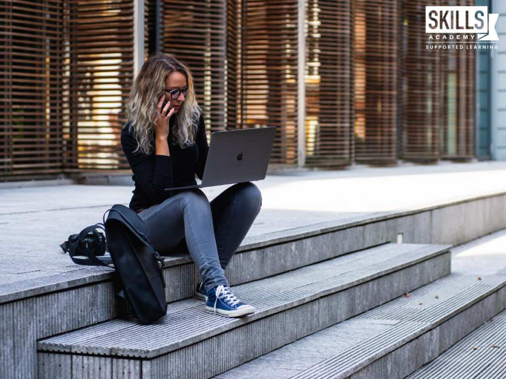Woman sitting on the steps outside of a building while talking on her cell phone with a laptop resting on her knee. Getting started with our Business English Course will help you become workplace ready.
