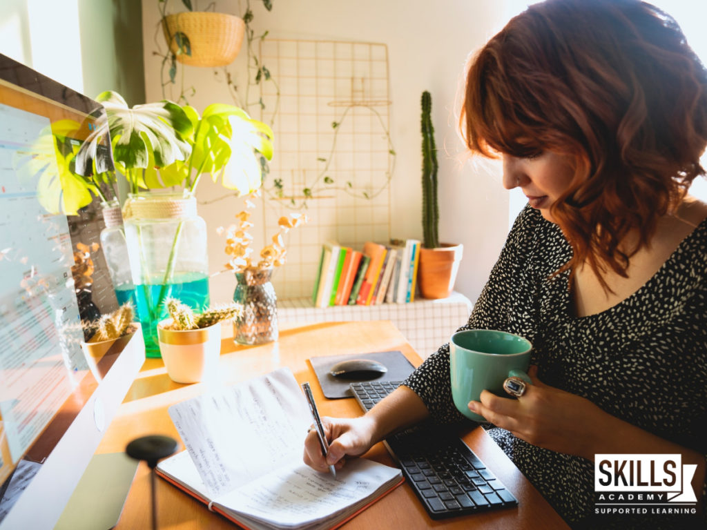 Woman writing notes in a book while studying at a desk in front of a computer and drinking coffee. Learn how to prepare your home for distance learning and create an effective study space.