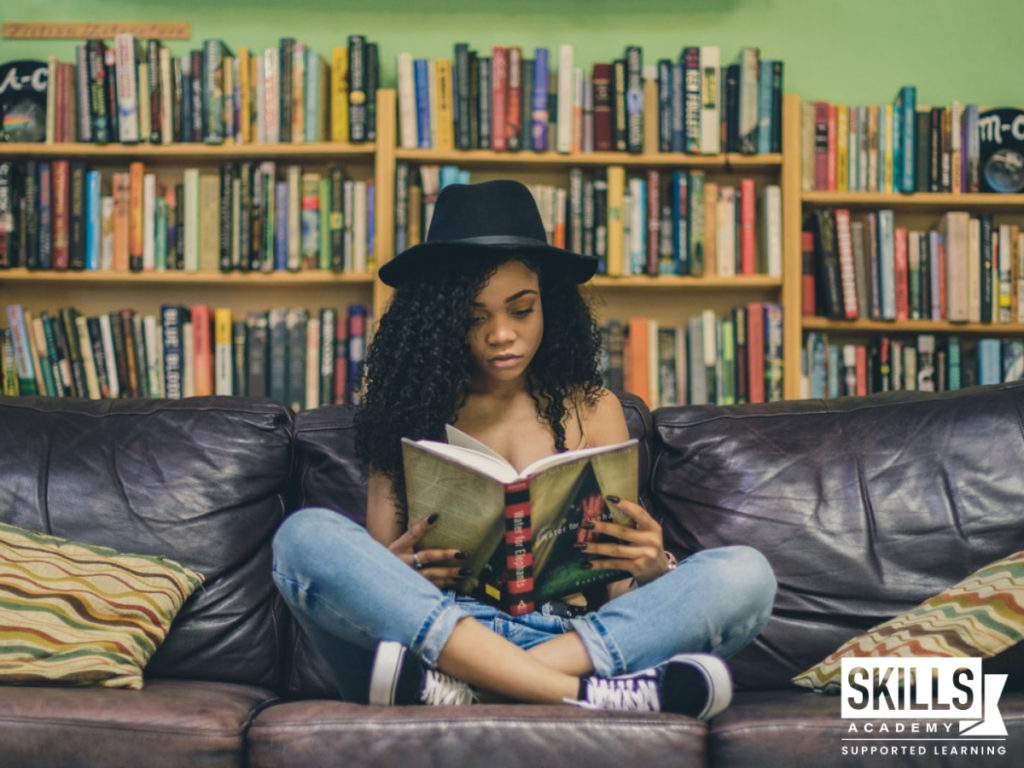 Woman sitting on a couch reading a book with bookshelves filled with books behind her. Find out what are your option after dropping out of high school are right here.