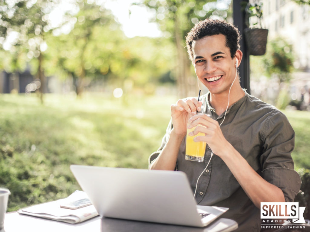 Guy smiling holding a glass of juice while sitting outside on a bench with a laptop in front of him. Join Skills Academy at any Time you want and you can study from anywhere you want.
