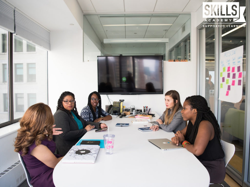 Team sitting around a table in an office having a meeting. Learn all about the skills every office professional needs right here.
