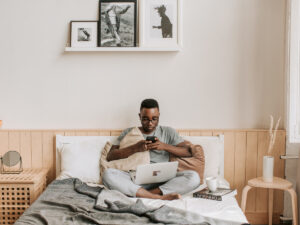 Guy sitting on his bed with his laptop on his lap and his cellphone in his hand. Questions to ask Before Accepting a Remote job.