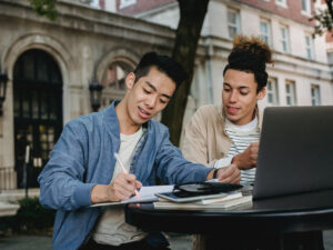 Two people writing down notes from a laptop. ICB Online Exam Rules.