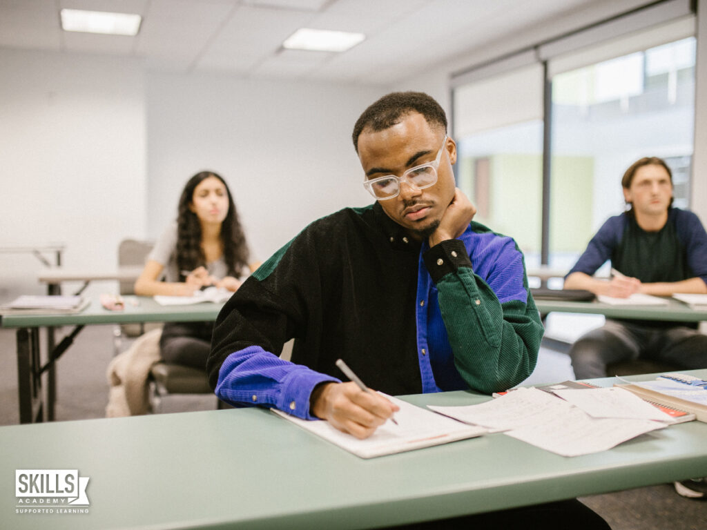 Student sitting at a desk in a classroom while writing on a paper. If you want tp pass your course, find out all about the ICB Pass Requirements right here.