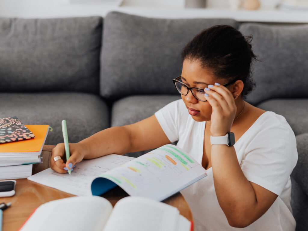 Student sitting on the floor at a coffee table, holding her glasses and making notes in her workbook. Study office administration courses online and update your skills.