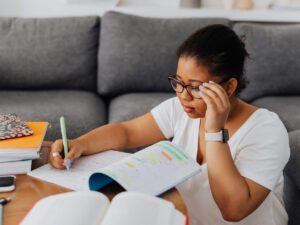 Student sitting on the floor at a coffee table, holding her glasses and making notes in her workbook. Study office administration courses online and update your skills.