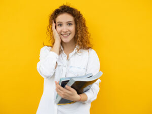 Woman holding her hand to her head while holding textbooks in her other hand, standing in front of a yellow wall. Explore what to do after studying ICB courses.