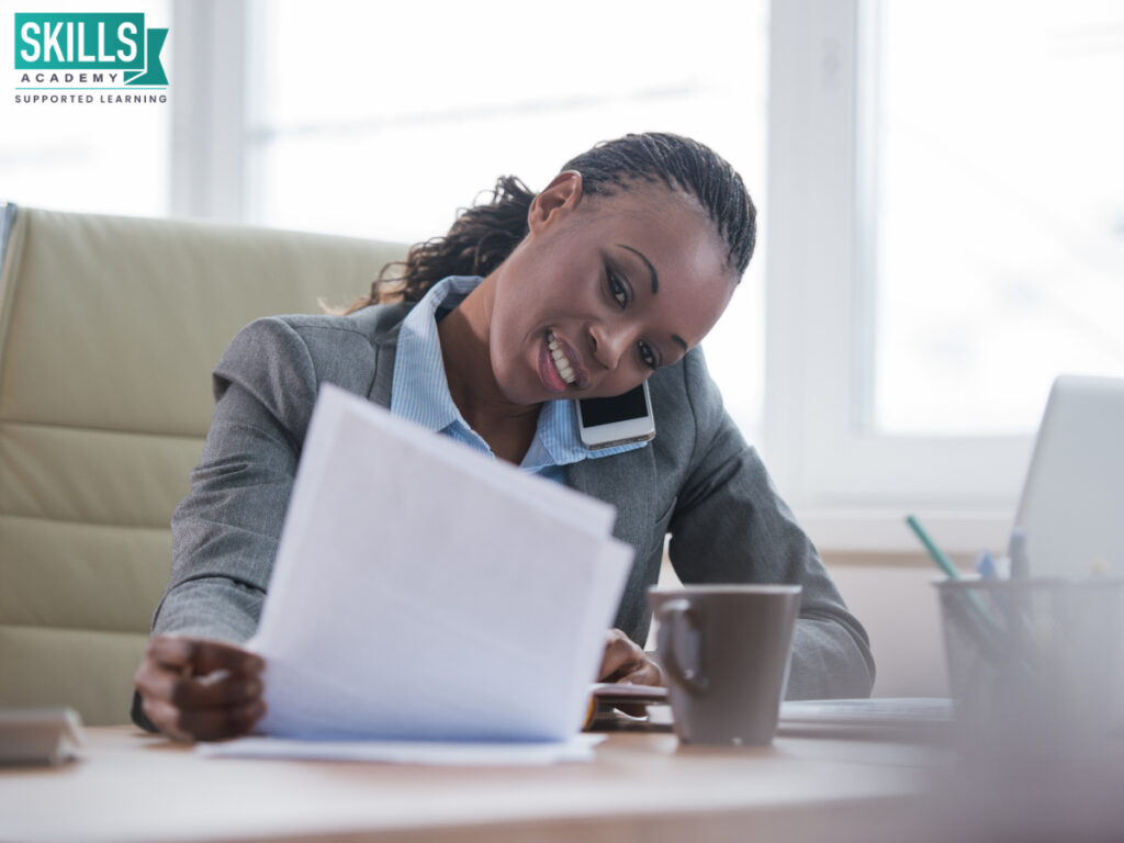 Office administrator sitting at a desk while talking on her phone and looking at sheets. Take a look at how to start a career in office administration.