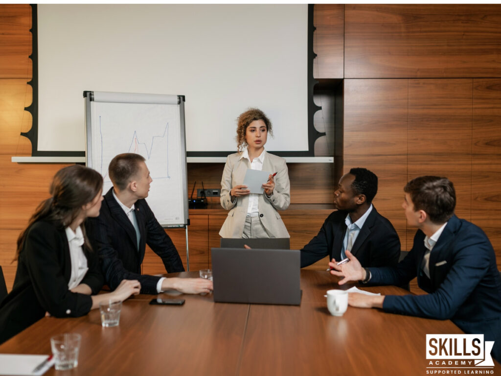 A young woman standing and leading a meeting at the office after studying our courses to help you become a business manager. Study with us today!