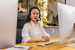 A young woman wearing a headset and sitting in front of a computer after learning all the skills every office administrator needs. Study our ICB Courses today!
