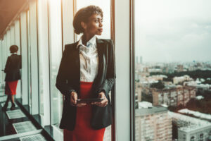 An African-American woman entrepreneur a digital tablet is leaning against a panoramic window of an office business skyscraper and looking in a distance; biracial businesswoman with a tablet pc. Do You Need Maths To Study ICB Business Management