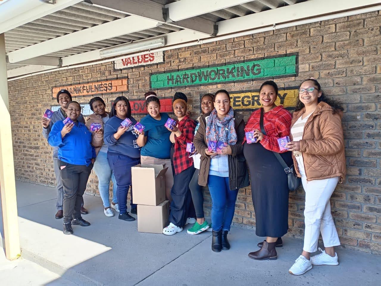 In the photo, our team stands proudly, holding up the donated pads in front of a mural painted with words like “Thankfulness” and “Integrity”—values that reflect who we are.